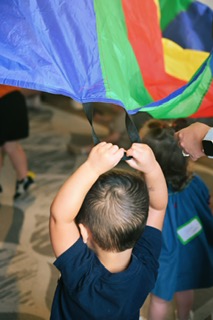 Children and grown-ups playing under a rainbow parachute in a Mommy & Me class